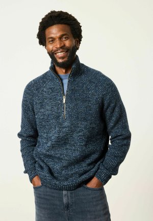 Mann mit Bart, der einen blauen Strick-Zip-Pullover und Jeans trägt, lächelnd mit den Händen in den Taschen vor einem schlichten hellen Hintergrund.