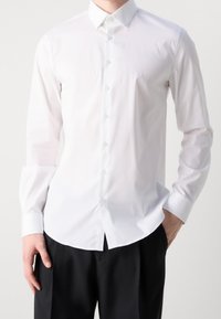 Man in een aansluitend wit overhemd met knopen, gestoken in zwarte broek, staand met één hand in zijn zak en de andere ontspannen.