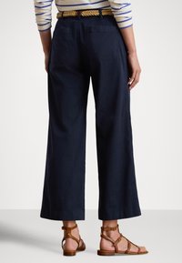 Tumši zili plaša stila bikses ar divām kabatām aizmugurē; aprīkotas ar dabīgu virves jostu. Kopā ar brūnām sandaletēm ar siksnām.