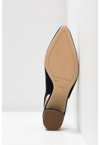 Semelle d'une chaussure à talon haut noire à bout pointu appuyée contre un coin blanc, indiquant la pointure 2½ et la marque Högl.