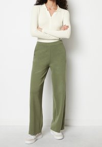 Pantaloni larghi di colore verde chiaro, realizzati in materiale morbido con vita elasticizzata, abbinati a un top a maniche lunghe color crema a coste e sneakers.