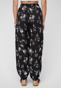 Femme portant un pantalon noir ample avec des motifs floraux roses et blancs, taille élastique et poignets aux chevilles, associé à des sandales sur un fond uni.
