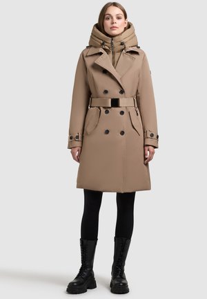 Manteau beige avec un col haut, des boutons double boutonnage, une taille ceinturée et des poches latérales. Porté avec des leggings noirs et des bottes de combat.