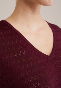 Bordeaux gebreid trui met een V-hals, voorzien van een zigzag kantpatroon en een getextureerde afwerking. Zacht materiaal met een lichte glans.