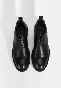 Zwarte leren schoenen met een gladde neuskap, een gestructureerd lichaam en een vetersluiting. Ze hebben een laag profiel en een rubberen zool.