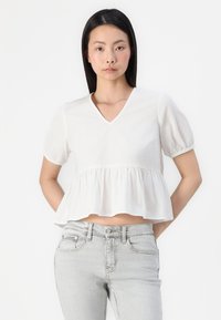 Fehér pamut blúz rövid puffos ujjakkal, V-kivágással és megkötött derékkal, fodros szegéllyel. Világosszürke farmert visel.