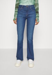 Jeans de campana azules hechos de denim, con cintura alta y sutiles detalles de costura, combinados con zapatillas blancas.