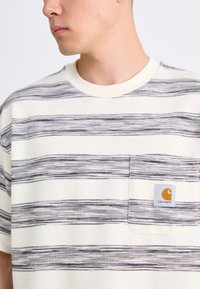 Stripete kortermet skjorte i kremfarge, med tynne marineblå linjer, en lomme på venstre bryst og en Carhartt-logo. Strukturt stoff.