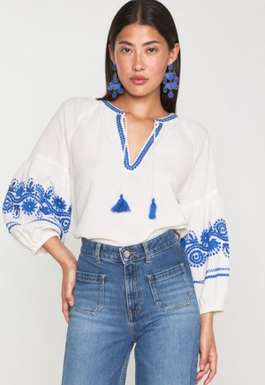 Weiße Bluse mit blauen bestickten Akzenten und Quasten Details an den Ärmeln, kombiniert mit hoch sitzenden Jeans aus mittlerem Denim.