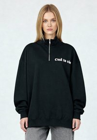 Sweat-shirt noir zippé à col montant, avec le texte blanc « C’est la vie » sur le devant. Coupe ample, poignets et ourlet côtelés.