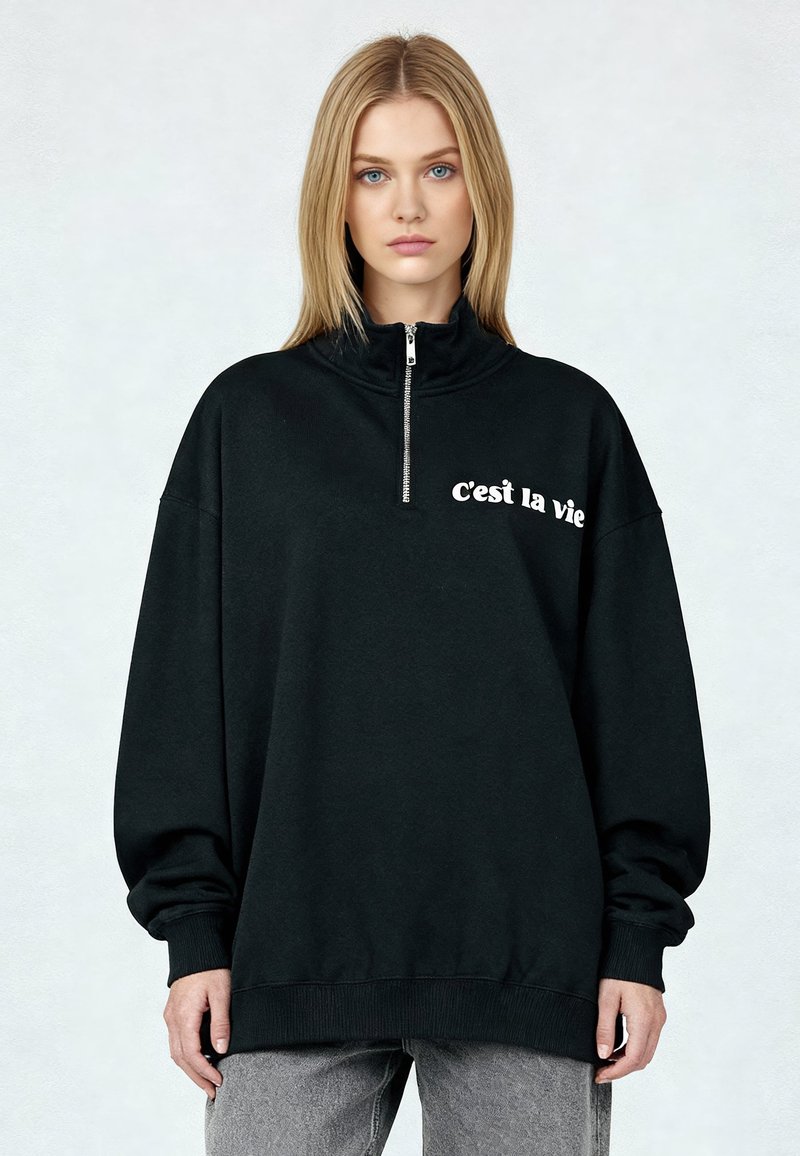 Sweat-shirt noir zippé à col montant, avec le texte blanc « C’est la vie » sur le devant. Coupe ample, poignets et ourlet côtelés.