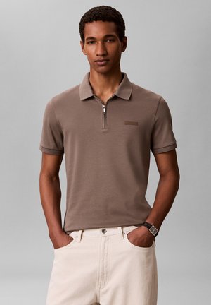Młody mężczyzna w brązowej polo z krótkim rękawem na zamek i białych spodniach, z rękami w kieszeniach, na neutralnym tle.