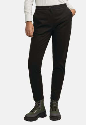 Person trägt schwarze, eng anliegende Hose mit Händen in den Taschen und grüne Schnür-Stiefeletten auf weißem Hintergrund.