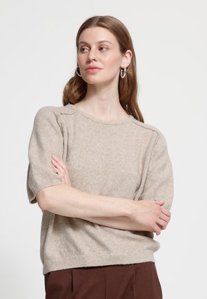 Femme aux cheveux châtain clair portant un pull beige à manches courtes et un pantalon marron, les bras croisés, regardant sur le côté avec des boucles d’oreilles en forme d’anneau.