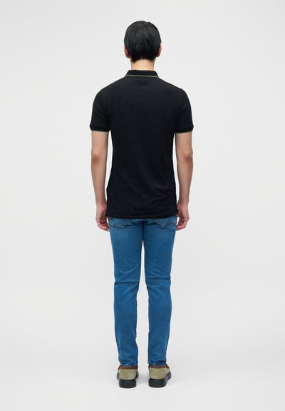 Μαύρο πόλο μπλουζάκι με πράσινη επένδυση, φορεμένο με μπλε τζιν παντελόνι και γκρι παπούτσια. Το μοντέλο φαίνεται από πίσω, με απλό φόντο.