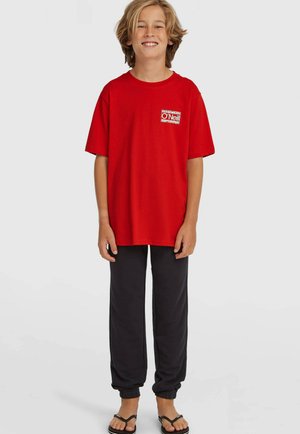 Rood katoenen T-shirt met korte mouwen en een klein logo op de borst, gecombineerd met donkergrijze joggingbroek en slippers.