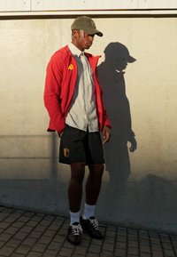 Athletic male wearing a red jacket, light gray shirt, black shorts with a logo, white socks, and black shoes, standing against a concrete wall.