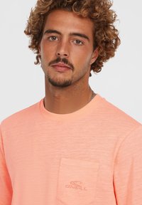 Kortærmet, koralfarvet T-shirt lavet af tekstureret stof med en lomme på venstre bryst, der har et broderet logo.