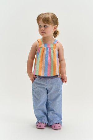 Kleinkind mit hellbraunen Haaren, das ein ärmelloses, regenbogenfarbig gestreiftes Oberteil, blaue Hosen und pinke Schuhe trägt und vor einem schlichten Hintergrund steht.