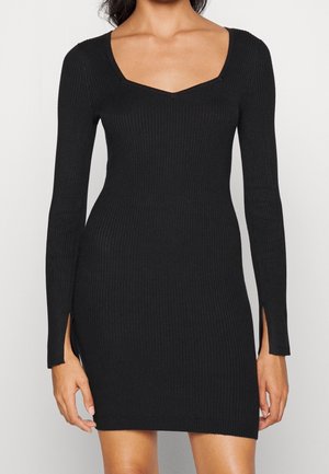 Kobieta ubrana w dopasowaną, długorękawową, czarną prążkowaną sukienkę z dekoltem w kształcie litery V i małymi rozcięciami przy mankietach.