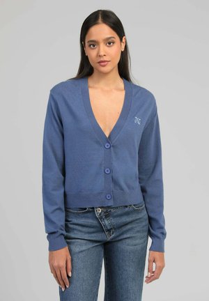 Jeune femme portant un cardigan bleu boutonné à col en V profond et un jean bleu, debout devant un fond gris clair uni.