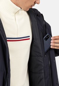 Černá bunda se zipovými kapsami, nošená přes bílý svetr s červenými a modrými pruhy. Telefon částečně zasunutý v vnitřní kapse.