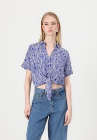 Kortermet, cropped skjorte i blå med hvit bølgedesign, med knappestolpe og knyttet nederkant. Brukt sammen med høytlivsblå jeans.