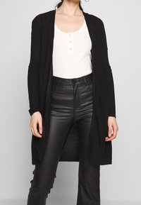 Cardigan noir à côtes avec ouverture frontale, manches longues et tissu texturé. Porté sur un top henley blanc et un pantalon noir ajusté.