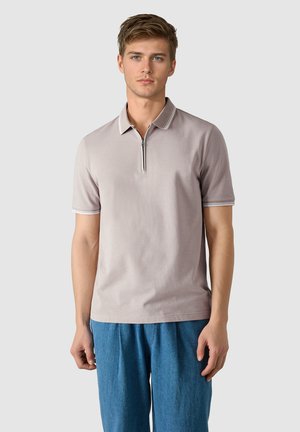 Ung mand iført en lysebeige kortærmet poloshirt med lynlås foran og blå afslappede bukser mod en ensfarvet baggrund.