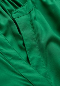 Tessuto verde con una superficie liscia e una leggera lucentezza. Presenta una cucitura e un design colletto piegato, che mette in evidenza un aspetto strutturato.