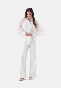 Blusa bianca con maniche a sbuffo trasparenti e un nodo al collo, abbinata a pantaloni bianchi a gamba larga e décolleté beige con punta. Tessuto dalla texture liscia.