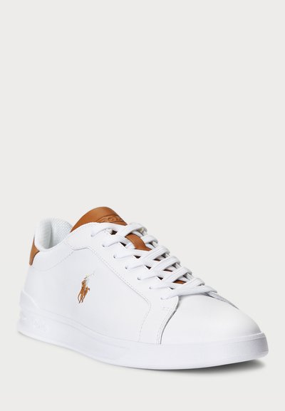 Hvite sneakers med en glatt læroverdel, brune lærdetaljer på hælen, og hvite snøringer. Har en liten brodert logo på siden.