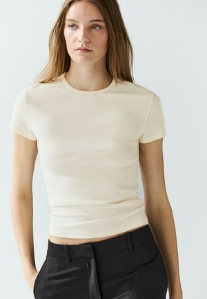 Jeune femme aux longs cheveux portant un haut beige clair uni à manches courtes et un pantalon noir, debout devant un fond clair uni.