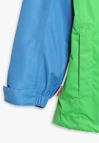 Giacca impermeabile blu e verde con una texture liscia. Presenta polsini elasticizzati, una tasca laterale e un'etichetta con dettaglio della bandiera.