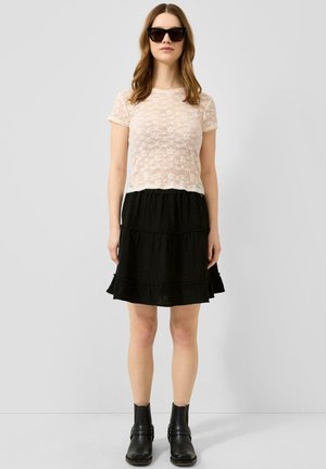 Femme portant des lunettes de soleil noires, un haut en dentelle transparente à manches courtes, une jupe noire à volants et des bottines noires, debout devant un fond clair uni.