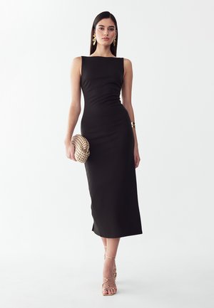 Frau trägt ein ärmelloses schwarzes Midikleid, goldene Blatt-Ohrringe, ein goldenes Armband, hält eine strukturierte goldene Clutch und goldene Riemchenabsätze.