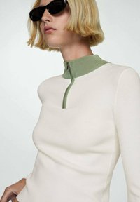 Långärmad topp i krämfärg med en grön dragkedjekrage. Mjuk textur med en åtsittande design. Bärs med svarta solglasögon, vilket ger en modern look.
