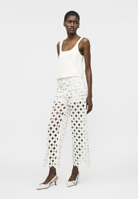 Femme portant un haut blanc sans manches, un pantalon large en crochet de dentelle, et des chaussures à talons blanches à bride arrière, debout devant un fond blanc uni.