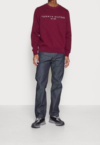 Bordowy sweatshirt z logo "TOMMY HILFIGER EST. 1985", w połączeniu z ciemnoniebieskimi dżinsami i czarnymi butami sportowymi z szarymi akcentami.