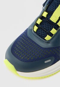 Chaussure de sport bleu marine avec tige en mesh, accents jaune vif, embout en caoutchouc texturé et semelle intermédiaire blanche. Dispose d'un système de laçage sécurisé.