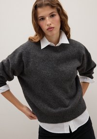 Knitpullover in Anthrazitgrau mit ribbelt gegen Ende und überschnittenen Schultern, getragen über einem weißen Hemd mit Kragen. Weiche Textur, lässige Passform.