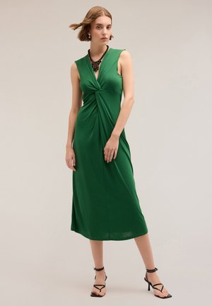 Grünes, ärmelloses Maxikleid mit einem verdrehten Vorderteil-Design, glattem Stoff und dezentem Glanz. Kombiniert mit schwarzen Sandalen mit Riemchen und einer Halskette.