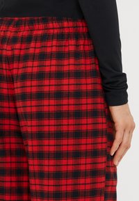 Røde og sorte ternede pyjamasbukser lavet af blødt stof med elastisk talje, der viser et nærbillede af mønsteret og kanten.