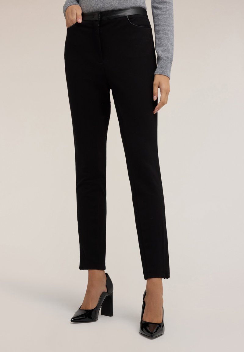 Zwarte broek van een gladde stof met een slim fit, voorzien van een ritssluiting en ceintuurhaken, gecombineerd met zwarte lakleren hoge hakken.