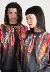 Two models wear black sweatshirts featuring multicolored abstract patterns in red, yellow, and white, emphasizing flowing shapes and designs.