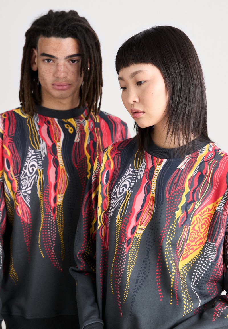 Twee modellen dragen zwarte sweaters met multicolor abstracte patronen in rood, geel en wit, die vloeiende vormen en ontwerpen benadrukken.