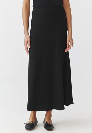 Jupe noire longue avec une texture lisse, design en A, et taille haute. Assortie à des chaussures plates noires.