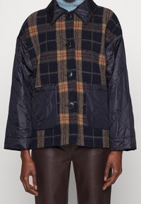 Rutig ull- och vadderad nylonjacka, med färgerna blå, brun och svart, två framfickor och knappstängning.