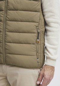 Olivgrüne Pufferweste mit horizontalen Nähten, Frontreißverschluss und einer Reißverschlusstasche mit farbigen Ziehlaschen. Getragen über einem hellen Pullover.