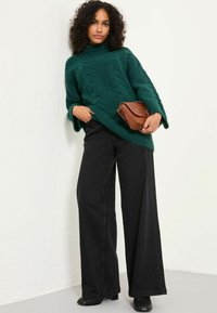 Grüner Kabelstrickpullover mit hohem Ausschnitt, kombiniert mit weiten schwarzen Jeans. Hält eine kleine braune Umhängetasche. Glatte Textur.